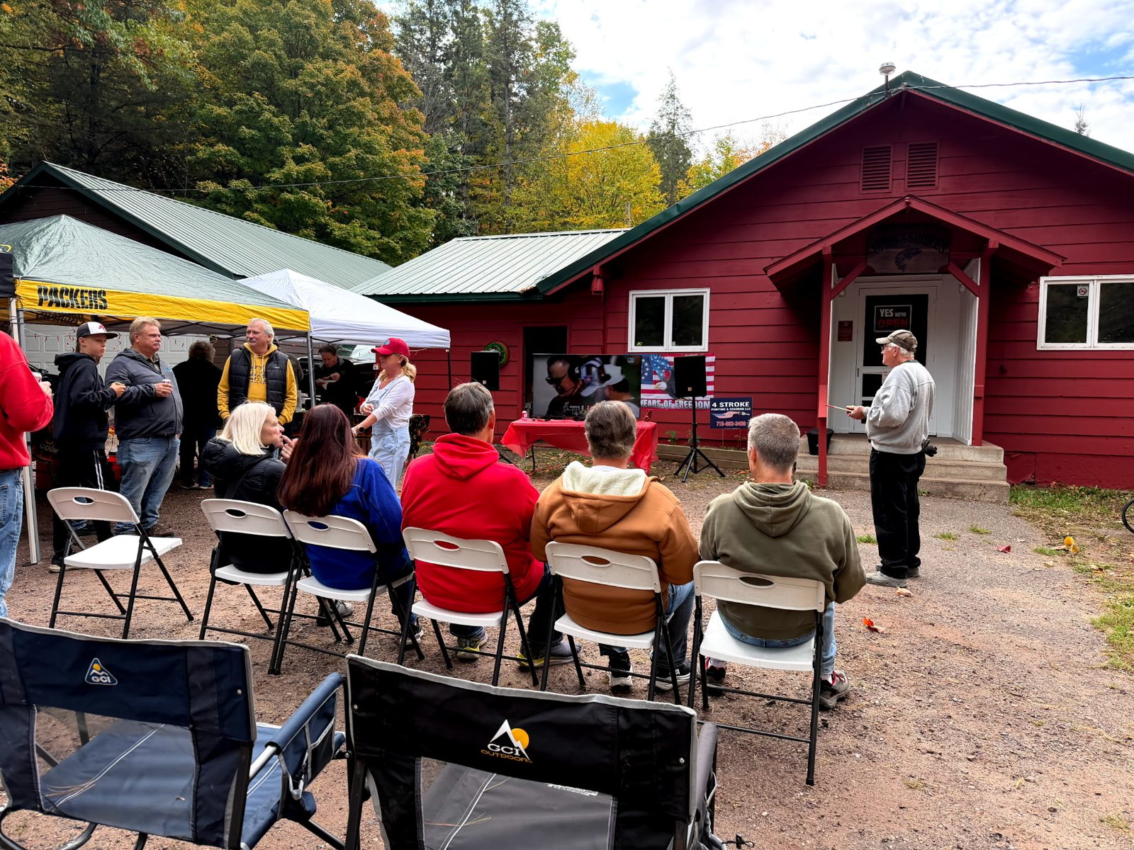 People sit in folding chairs outside a red building with a white awning.