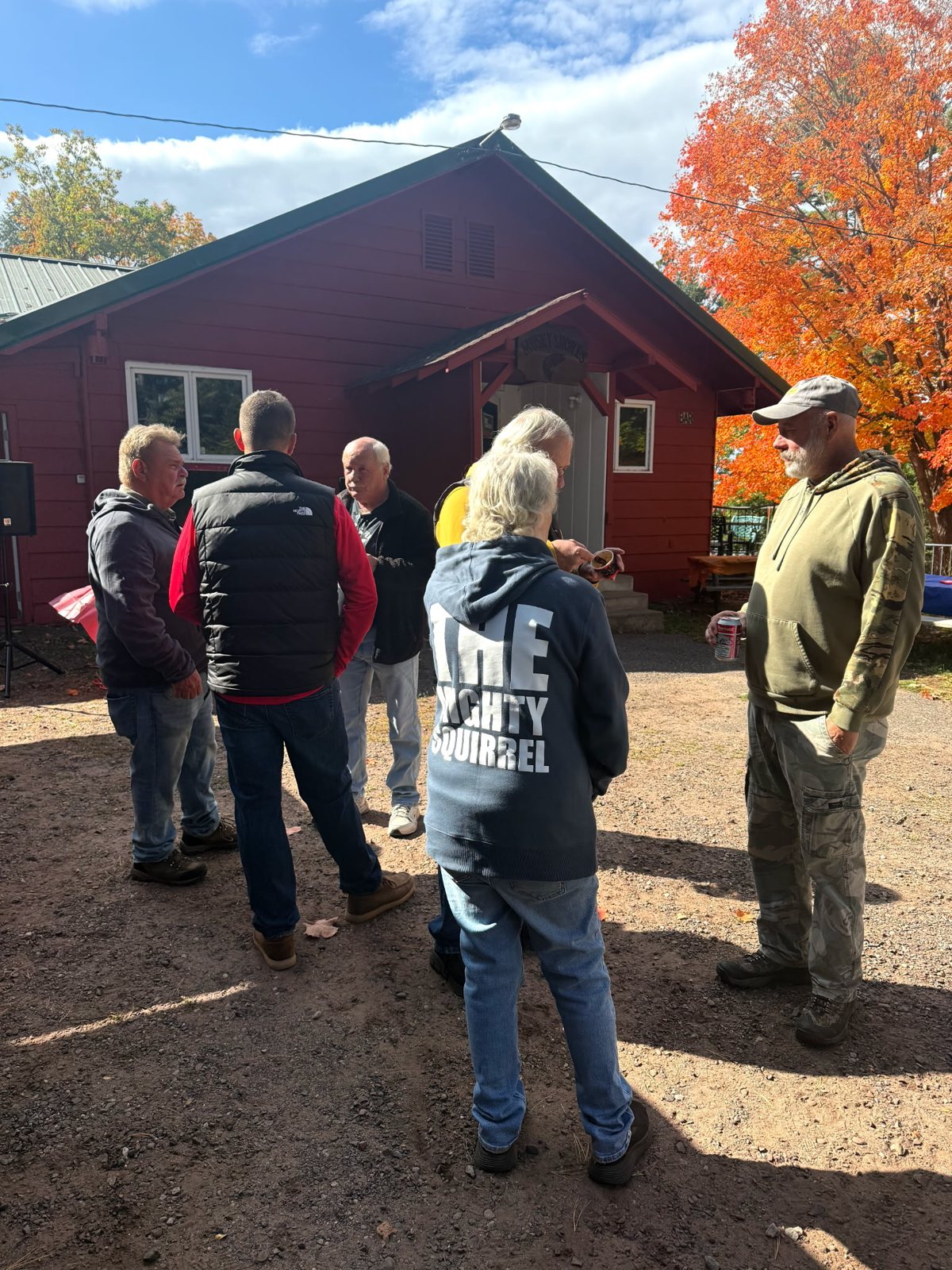 A group of people talk in the yard outside the red building, beside a tree in fall color.