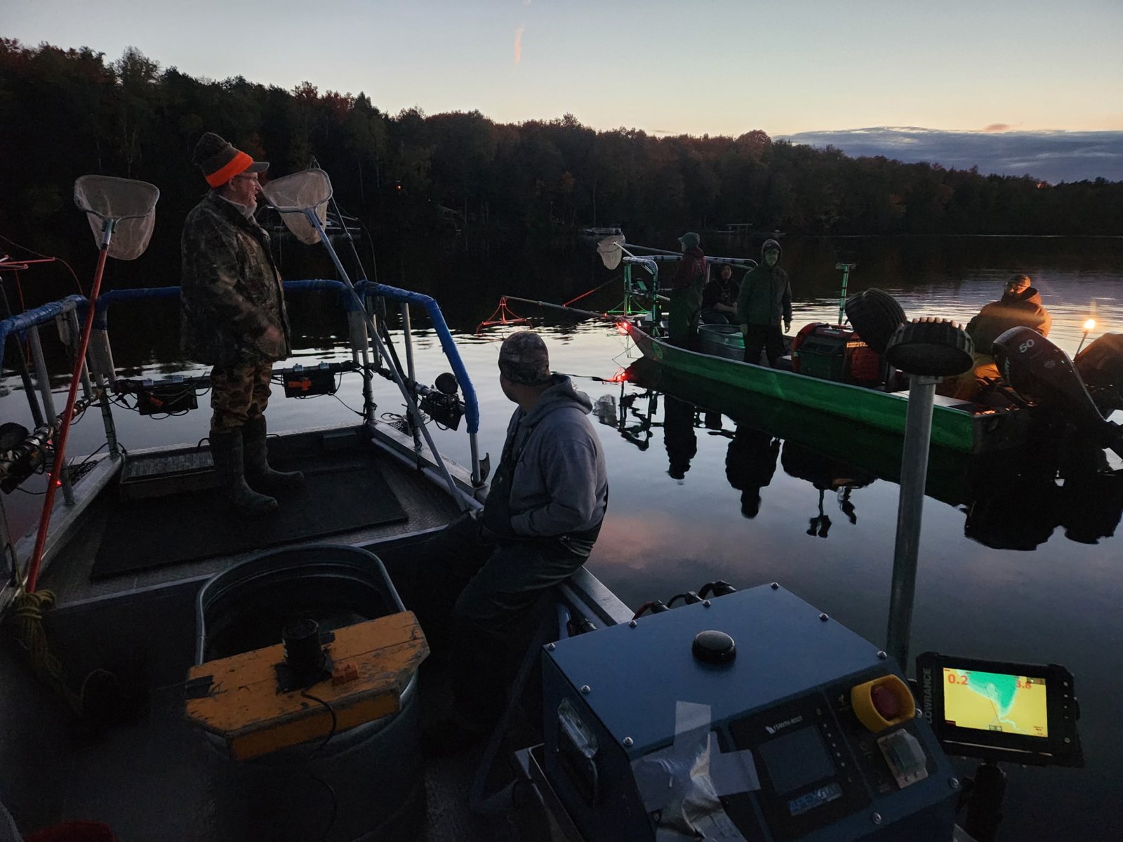 Two boats on calm water at dawn, with wooded shore behind.