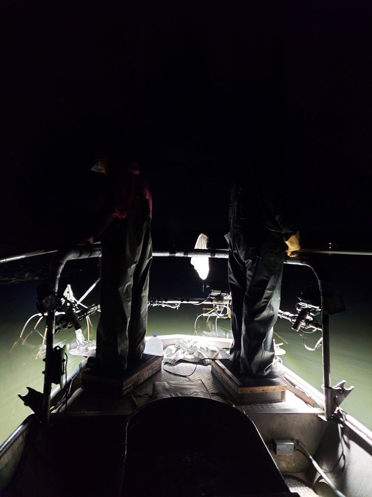 Two crew members stand at the bow of the survey boat under its lights at night.