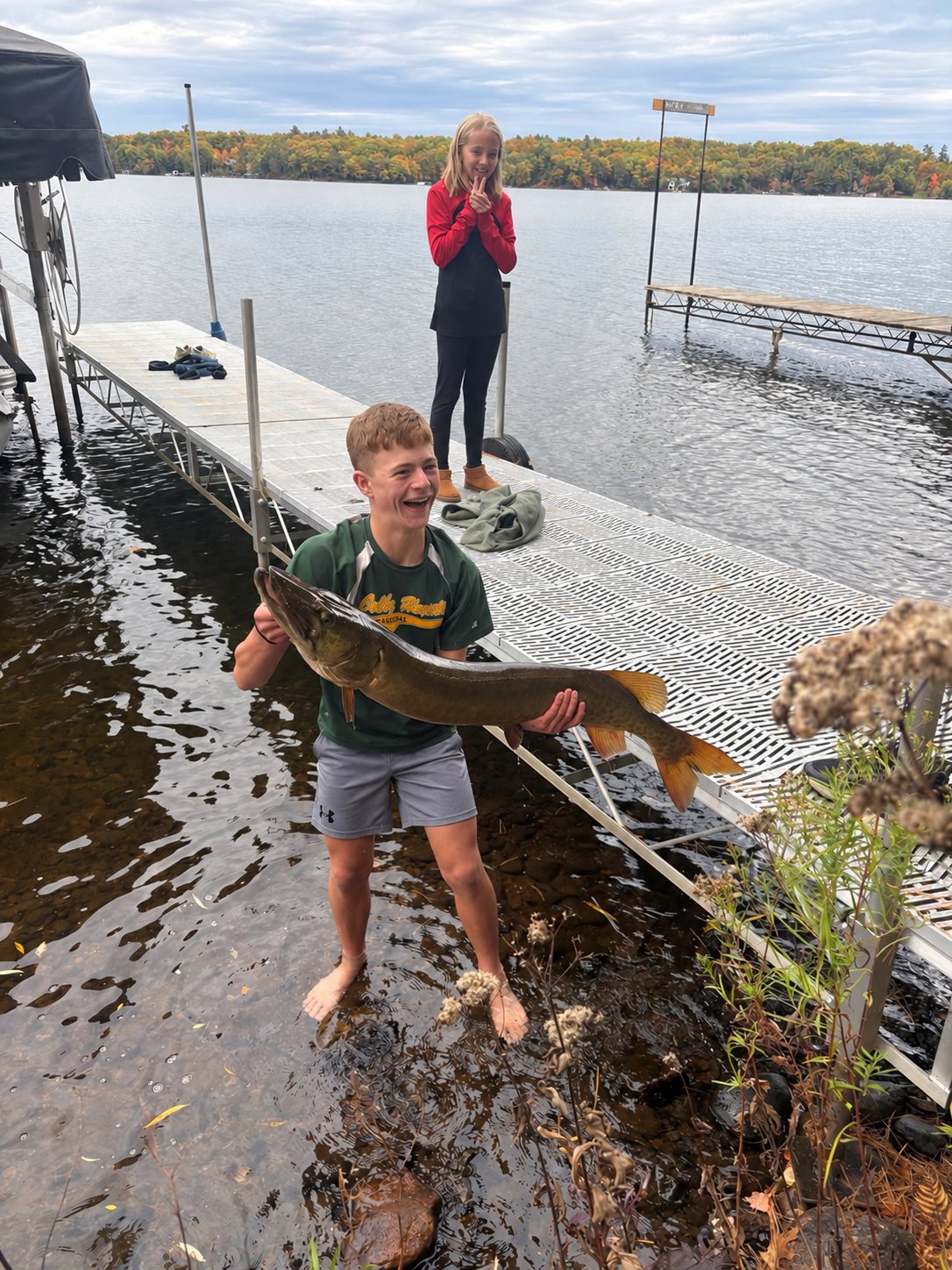 A young angler standing in shallow water beside a dock holds up a large muskellunge, with fall colors on the far shore.