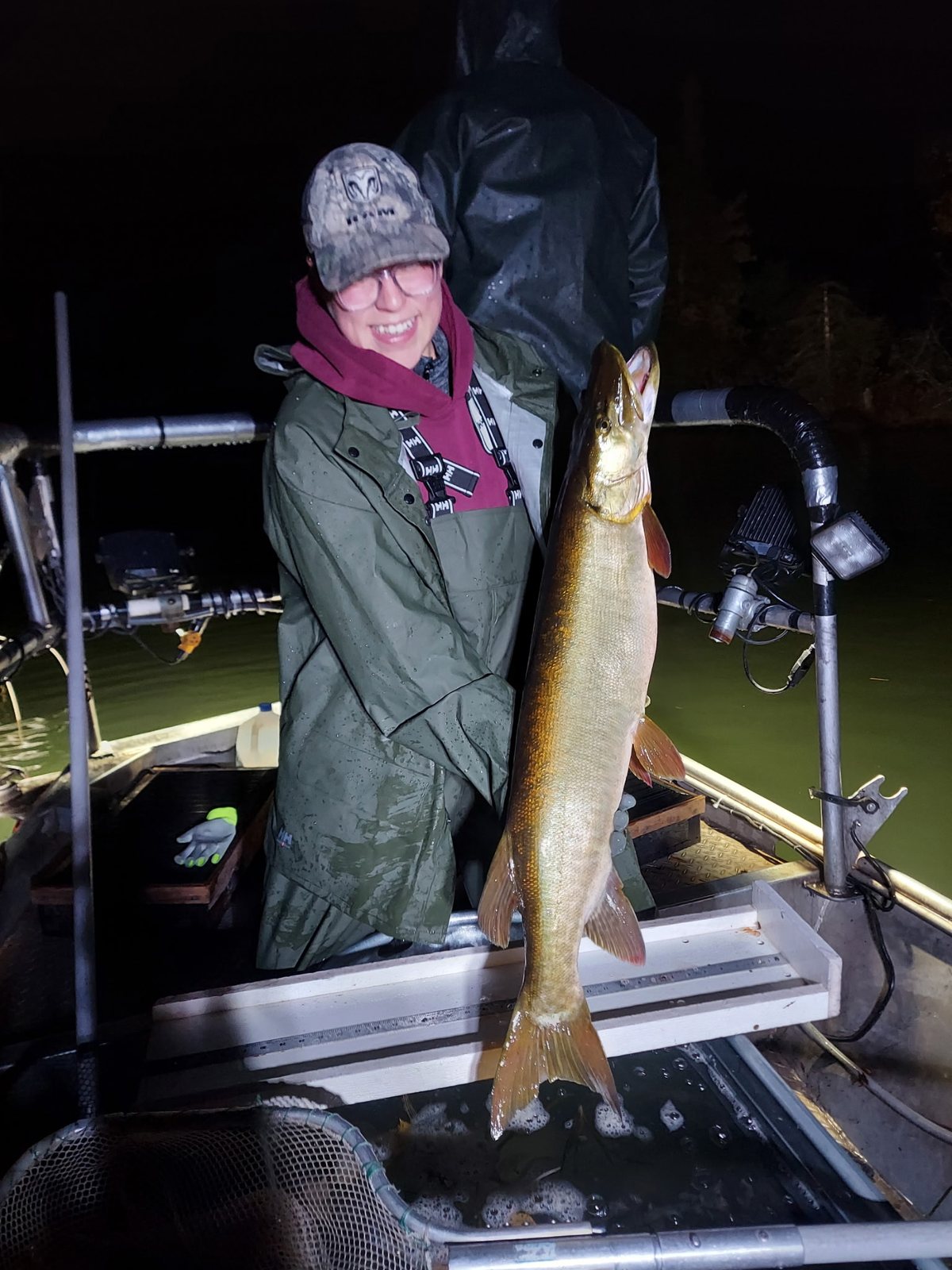 A survey crew member in rain gear holds up a muskellunge on a boat at night.